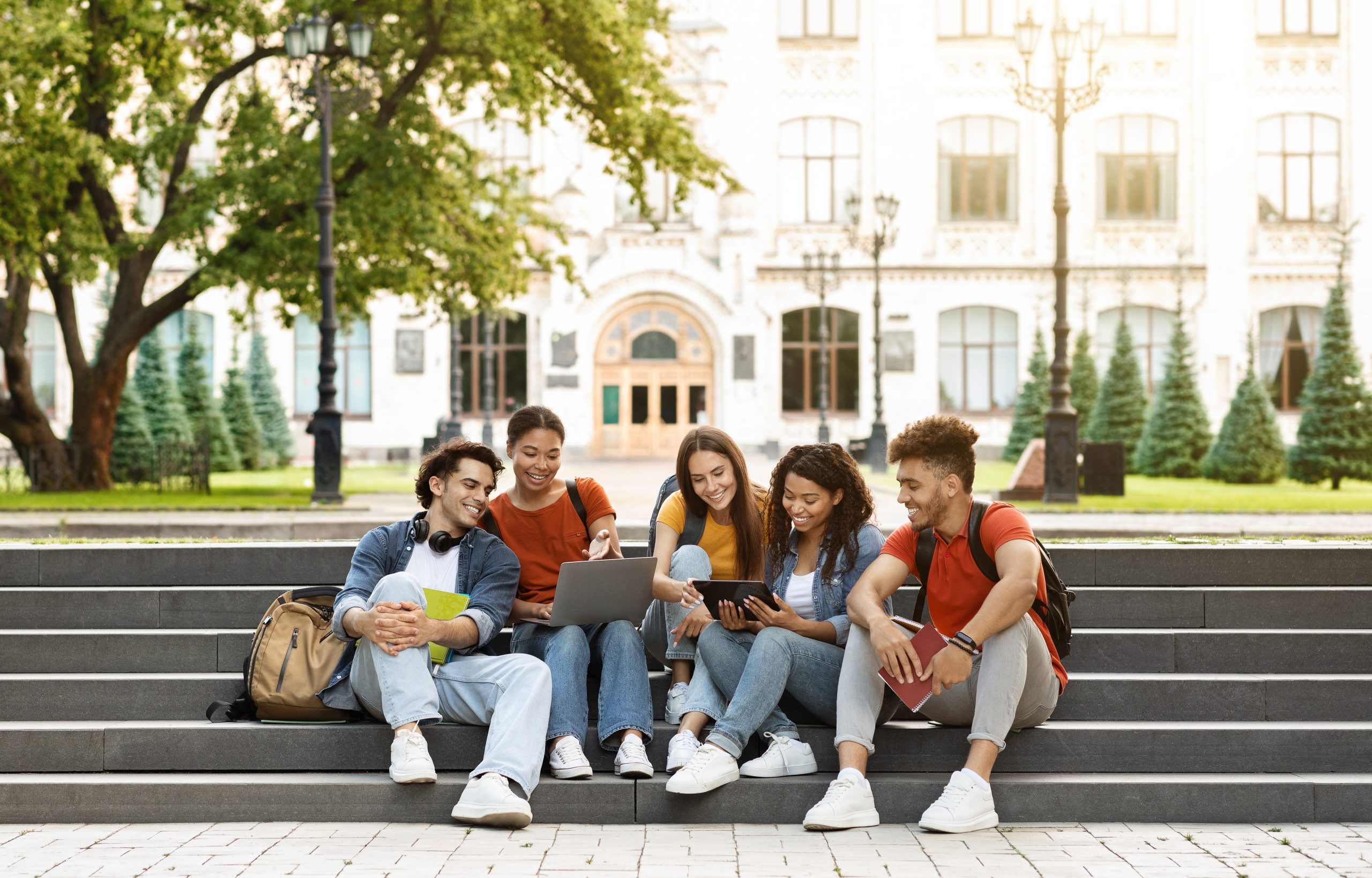 Group Of Students With Digital Tablet And Laptop Study Together Outdoors, Happy Multiethnic Young Friends Sitting On Stairs Near University Building, Using Modern Gadgets For Education